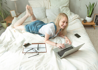 Beautiful happy girl in denim jumpsuit is lying on a white bed with laptop.