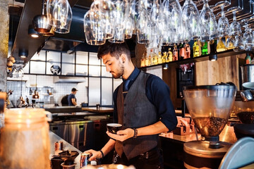 A young Caucasian barista pours milk into coffee espresso, making cappuccino or latte. A professional barista preparing coffee with inspiration on the bar counter