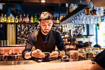 Professional bartender waiter with smile serving coffee to young woman. Regular visitor, sitting at the bar, communicating with interest with the barista. In favorite cafe with pleasant, yellow light