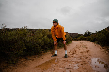 Tired young male trail runner resting after sprinting down wet mountain trail in cloudy weather with rain and foliage