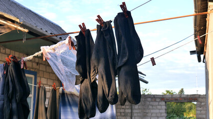 Several pairs of black socks dry on a clothesline next to other laundry and a washed plastic bag in the yard of a peasant house.