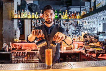 Handsome brunette barista of East Caucasian appearance preparing a drink at the bar in a restaurant. Mixing a coffee and orange juice by adding ice to the cocktail. Inserts a drinking straw