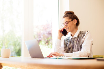 Happy young woman making a phone call while sitting at desk and working on laptop online