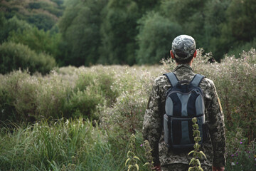 A man in a camouflage uniform with a backpack behind his back stands against the background of the forest
