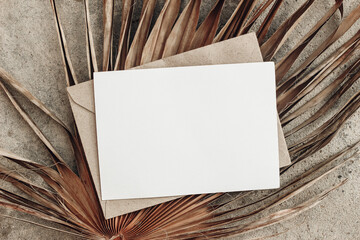 Summer stationery still life. Closeup of blank card mock-up and craft envelope on dry palm leaf. Grunge beige concrete background. Flat lay, top view. Tropical vacation concept. Moody boho design. © tabitazn