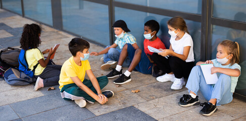 Group of preteen children in face masks resting outdoors in schoolyard during break in lessons. Back school concept during pandemic.