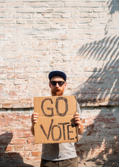 Fototapeta na wymiar Man shows cardboard with Go Vote sign on brick wall urban background. Voting concept. Make the political choice, use your voice. Bearded guy in sunglasses invite to go to the presidential elections.