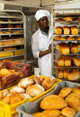 Experienced African American baker working in bakehouse, pushing rack trolley with freshly baked loaves