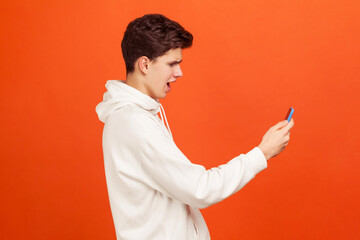 Profile portrait of shocked teenager in white casual sweatshirt looking at screen of mobile phone with excited face, receiving email with sale coupons. Indoor studio shot isolated on orange background