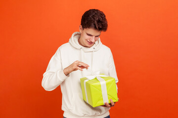 Handsome young man in casual white sweatshirt with hood adjusting bow on giftbox and smiling,...