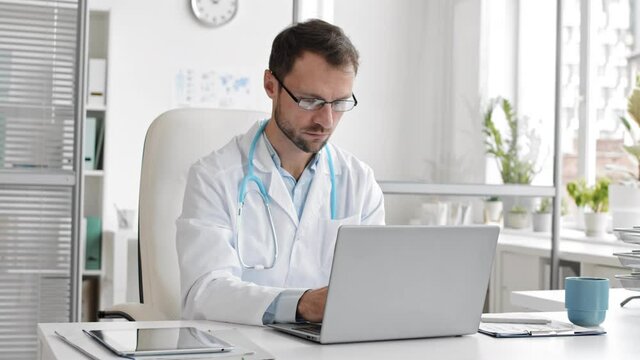 Panning of middle-aged Caucasian man wearing medical gown and glasses is sitting at desktop in the office and working on PC