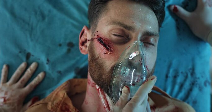 Close up of paramedics hands putting on oxygen inhalation mask on bearded man face. Unconscious guy in 40s with head traumas lying on hospital stretcher in ambulance