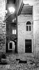 Fototapeta na wymiar A narrow deserted street in the medieval historical part of Trogir, Croatia, lit by lanterns at night in black and white tones