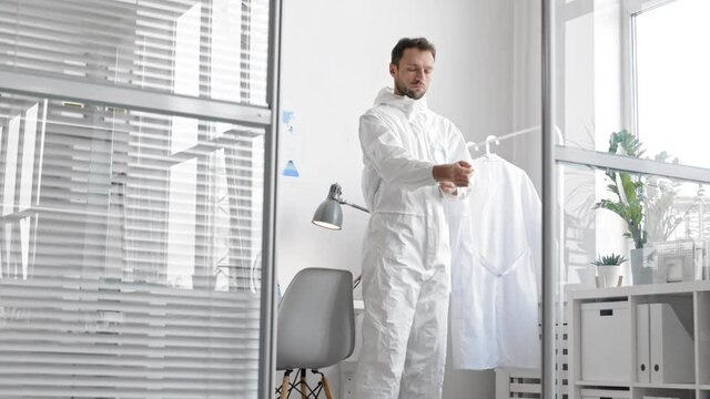 Medium shot of middle-aged Caucasian male doctor is standing in the office, zipping up white protective coverall and then going away