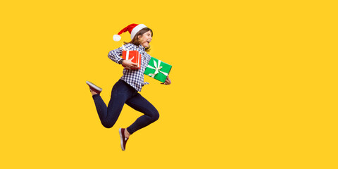 Side view portrait of extremely joyous brunette woman in santa hat jumping or running in air with wrapped xmas gift boxes, celebrating discounts. indoor studio shot isolated on yellow background