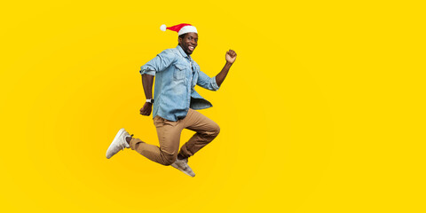 Fototapeta na wymiar Full length portrait of happy joyous man with new year santa hat jumping or flying, hurry running to his dream, looking at camera with toothy smile. indoor studio shot isolated on yellow background