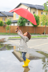 Happy toddler girl playing with a red umbrella in the street near the house. Kid in yellow rubber rain boots in a puddle