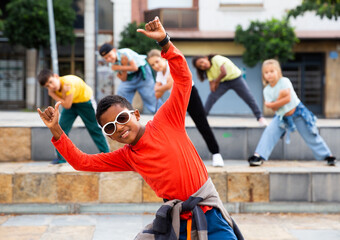 Portrait of confident african boy in sunglasses dancing hip-hop with group of tweens on summer city...