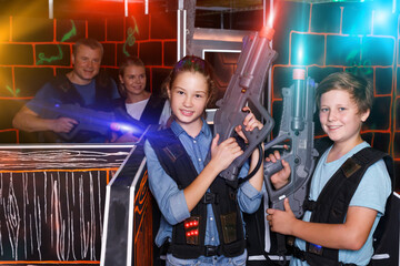 Ordinary teenage boy and girl posing with laser guns while having fun with adults on laser tag arena