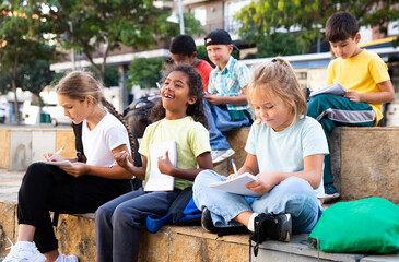 Smiling kids sitting on bench and writing in notepads, studying outdoors