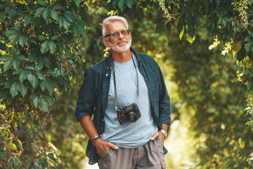 A confident elderly man walks with a camera in a city park.