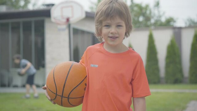 Little boy looking at camera and smiling as blurred kid playing basketball at the background. Portrait of cute Caucasian child posing outdoors with ball. Sportive childhood.