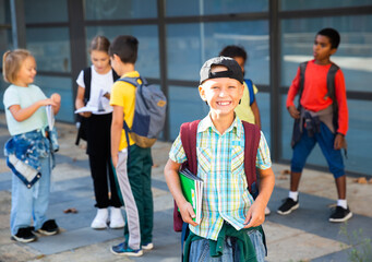 Cute cheerful tween boy looking at camera while standing in school campus with notebooks in hands on background with schoolmates.