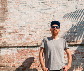 Handsome hipster man with lush beard and mustache on light brick wall background palm leaves shadows. Red bearded guy in cap black sunglasses outdoor.Serious brutal male stylish macho photo copy space