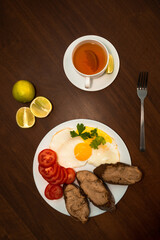 Breakfast on a plate of fried eggs with sliced tomatoes, herbs, slices of gray bread with chicken pate and tea with lime slices on a dark brown wooden table