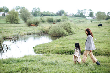 Stylish mother and daughter have fun outdoors in a field with green grass by the river when it rains