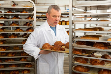Portrait of positive baker working in bakehouse, putting tray with fresh bakery goods on rack trolley..