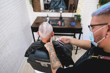 Hipster senior man getting hair cut with a razor and clipper in vintage barbershop - Barber wearing protective mask for coronavirus spread prevention - Focus on customer