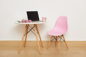 A pink chair and a white table against the wall. On the table are a laptop, glasses, a cup and a bouquet of roses. Studio interior or workplace.