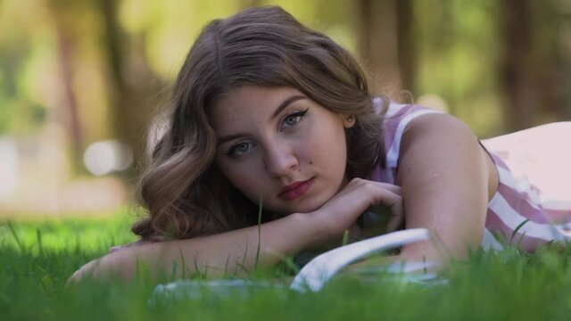 Young woman with curly fair hair rests lying on green grass near opened thick book and looks straight leaning on hand in park on summer day, slow motion closeup