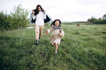 Stylish mother and daughter have fun outdoors in a field with green grass by the river when it rains