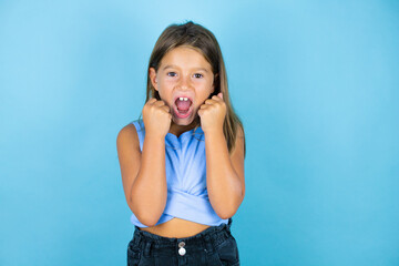 Young beautiful child girl over isolated blue background very happy and excited making winner gesture with raised arms, smiling and screaming for success.