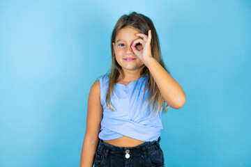 Young beautiful child girl over isolated blue background doing ok gesture shocked with smiling face, eye looking through fingers