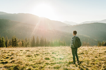 Young tourist man with a backpack on top of enjoying the sunset. Back view. Hipster walks in the autumn grass in the mountains on vacation. Holiday trip concept. World Tourism Day.