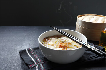 a bowl of hot vegetarian ramen noodle is on the black table with a dim sum basket and bottle of olive oil during lunch in the Chinese restaurant