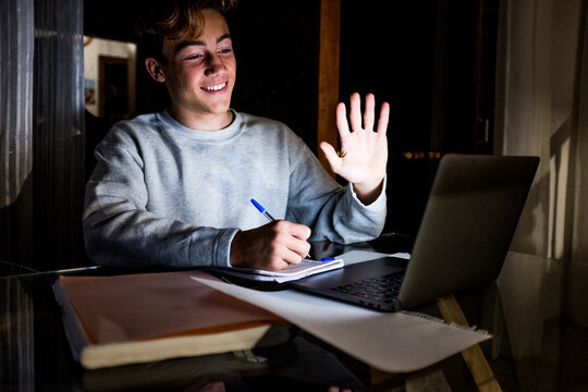 close up and portrait of young man or teeager waving at someone in video call doing homeworks and smiling looking at the computer pc or laptop