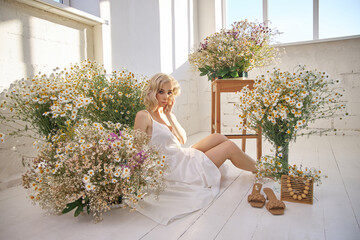 Beautiful blonde woman in a white dress is sitting on the floor among the chamomile flowers....