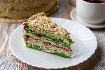 Slice of green pistachio sponge on a wooden table, with a whole torte on background