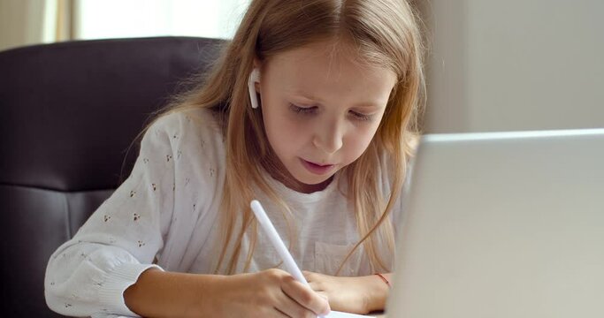 Distance learning concept. Active schoolgirl studying online using laptop making notes in copybook. Teen lady wearing headphones, looking in web cam, wave hands in greeting, sitting at home desk