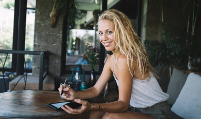 Portrait of cheerful blonde female in casual wear sitting in cafe interior making notes in notepad,...