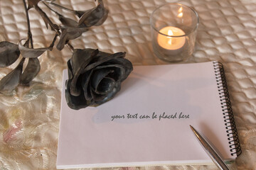 Hygge style. a lighted candle illuminates a notebook on which a silver pen lies. a forged rose, as a symbol of eternal love, lies nearby. vintage template, style messages
