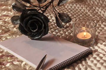 Hygge style. a lighted candle illuminates a notebook on which a silver pen lies. a forged rose, as a symbol of eternal love, lies nearby. vintage template, style messages
