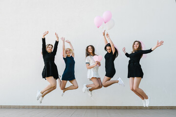 Bride and bridesmaids jumping near white wall outdoors and celebrating bachelorette party. Beautiful sexy young girls in dress and veil on bachelor party. Maiden evening, Hen-parties. Place for text.