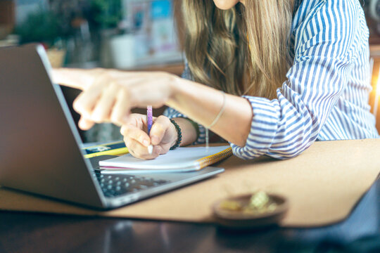Business woman writes in notebook on desk and pointing hand at computer screen. Girl working from home office kithcen. Using laptop and online shop. Distance learning online education and work