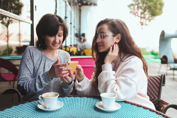 Mature mother and her young daughter sit together in cafe or restaurant. Girl shows her mother something on yellow smartphone. Watching together and having fun. Using modern technologies.