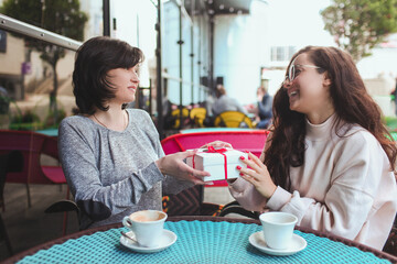 Mature mother and her young daughter sit together in cafe or restaurant. Girl give her mom present. White box with red stripes. Gift or surprise for mature mom. Enjoy drinking coffee outside.